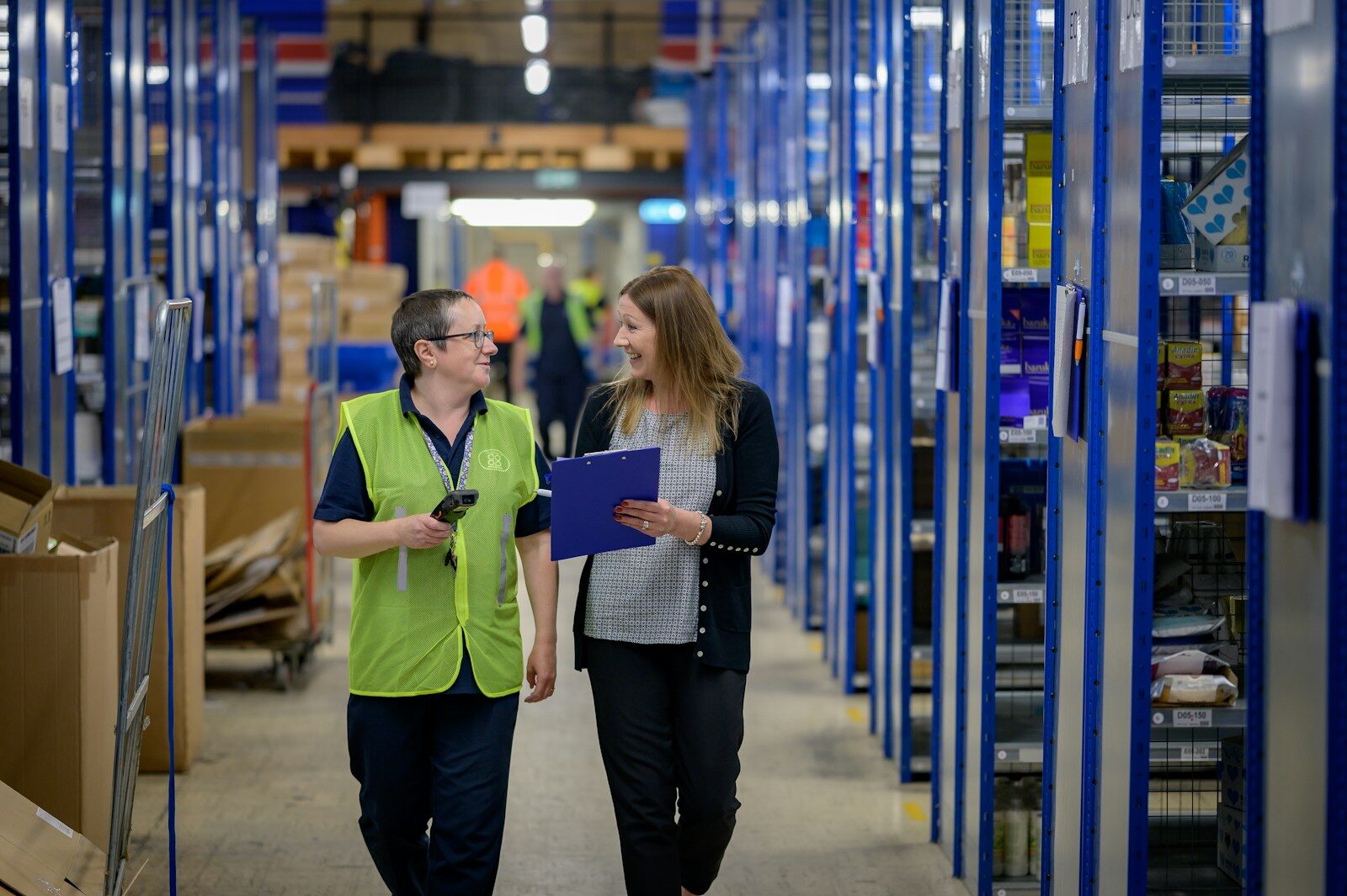 Two women walking through a warehouse aisle with blue shelving units